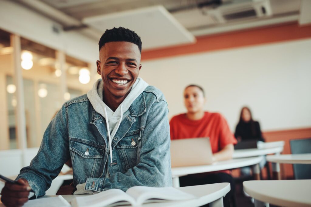 Happy teenage boy in a denim jacket smiling in a classroom, representing teens who get braces from Dr. Matthew Noyce at Noyce Orthodontics in St. Joseph & Maryville, MO.