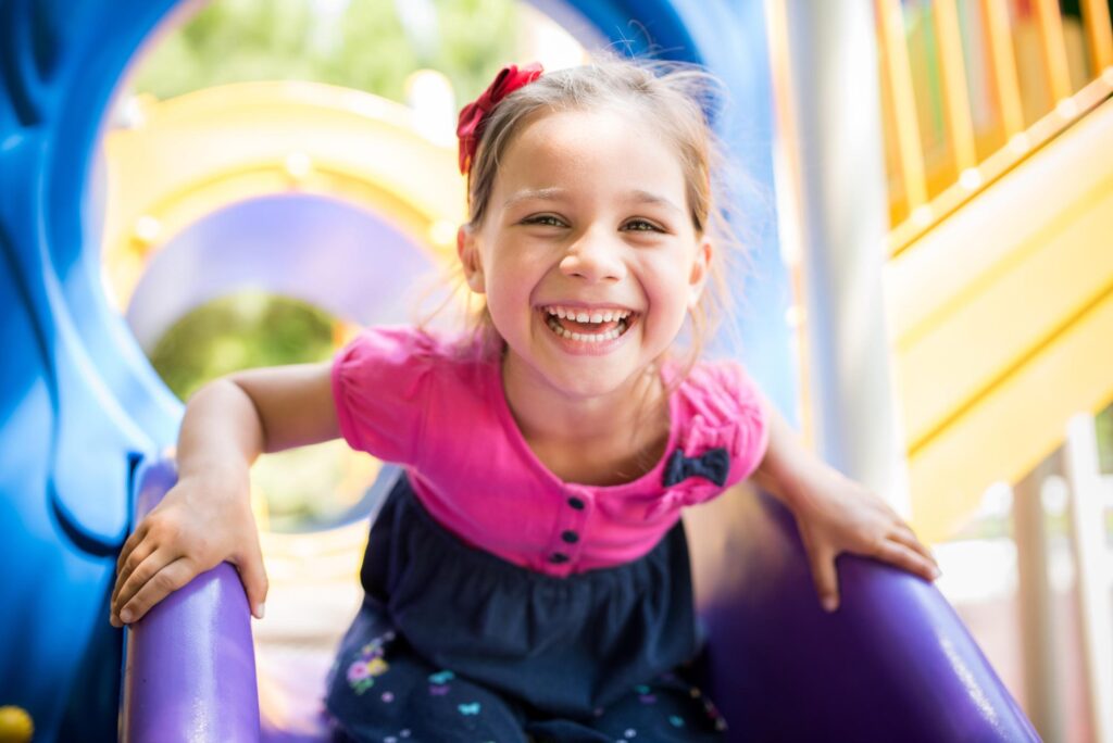 Happy young girl in a pink shirt with a red bow in her hair, laughing on a colorful playground, showing the confident smiles Dr. Matthew Noyce helps kids get at Noyce Orthodontics.