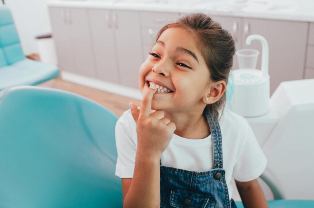 Young girl in a white shirt and denim overalls smiling in a dental chair at Noyce Orthodontics, where Dr. Matthew Noyce treats children in St. Joseph and Maryville, MO.